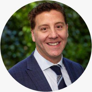 Man wearing a coat and tie, smiling against a background of greenery